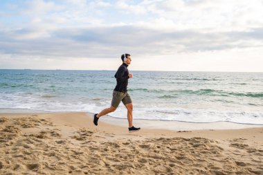 Fit male runner training on the summer beach and listen to music against beautidul sky and sea