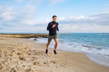 Fit male runner training on the summer beach and listen to music against beautidul sky and sea