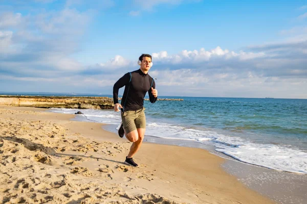 Fit male runner training on the summer beach and listen to music against beautidul sky and sea