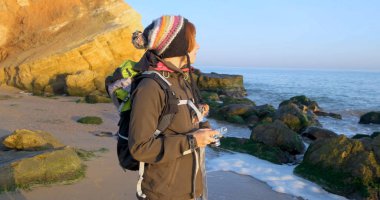 Sırt çantası ve retro film kamerasıyla genç bayan gezgin deniz kenarındaki sonbahar dağlarında seyahat ediyor.