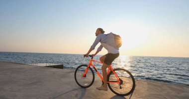 Genç ve yakışıklı bir erkek, günbatımına ve denize karşı sabah sahilinde renkli bisiklete biniyor.