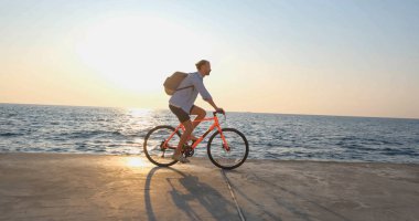 Genç ve yakışıklı bir erkek, günbatımına ve denize karşı sabah sahilinde renkli bisiklete biniyor.