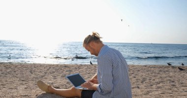 Yaz plajında genç yakışıklı erkek güneş doğarken dizüstü bilgisayarla çalışıyor.