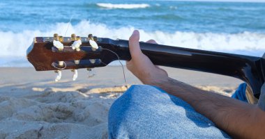 Genç yakışıklı erkek, güneşli bir günde sahilde akustik gitar çalar. Arka planda deniz ya da okyanus 4k Uhd.