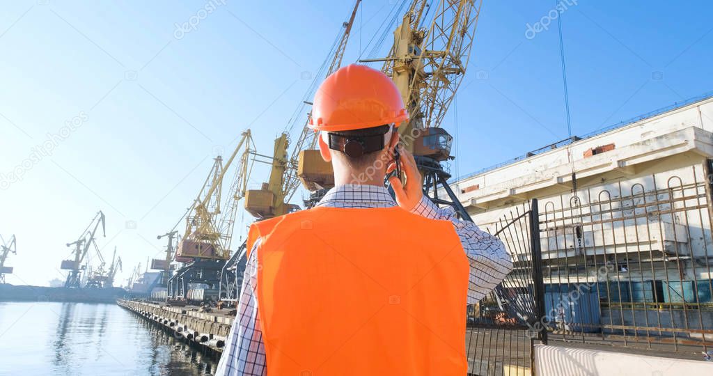 male worker of sea harbor in orange helmet and safety west, cranes and sea background 