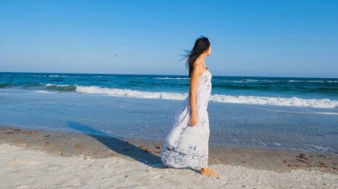 Beyaz elbiseli güzel bir kadın yaz plajında yalınayak yürüyor.