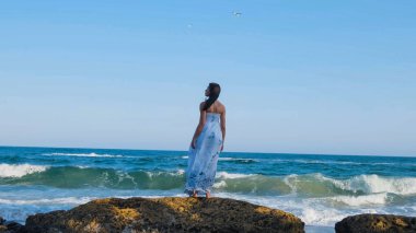 Beyaz elbiseli güzel bir kadın yaz plajında yalınayak yürüyor.