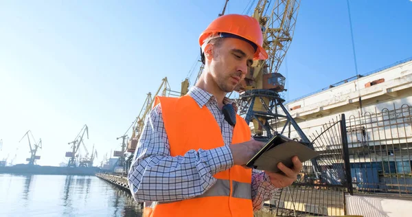 male worker of sea harbor in orange helmet and safety west, cranes and sea background