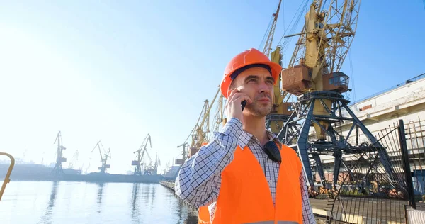 Turuncu miğfer ve güvenli batı, vinçler ve deniz altyapısı ile deniz limanında çalışan erkek işçiler. 