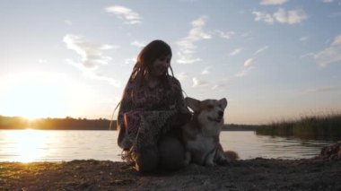 Young happy hipster woman having fun with corgi dog on the beach against sunset