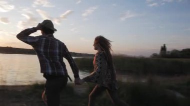 Young happy couple of lovers hiking near river in summer time against beautiful sunset