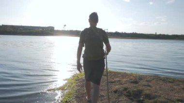 Young male with backpack and wooden hiking staff crossing a big river in summer sunny day