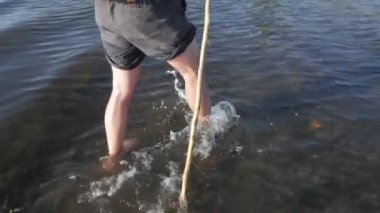 Young male with backpack and wooden hiking staff crossing a big river in summer sunny day