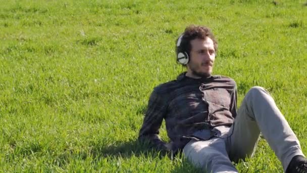年轻的雄性在绿草上悠闲自在地躺在夏天的田野里 用白色的大耳机听音乐 — 图库视频影像
