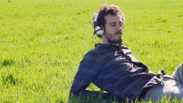 緑の芝生でリラックスして白いヘッドフォンで音楽を聴いたり夏の畑に寝そべったり — ストック動画