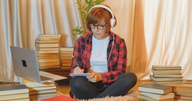 Evde birçok kitap ve dizüstü bilgisayarla okuyan genç bir kız öğrenci.