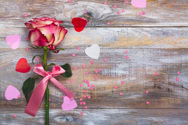 Ροζ και κίτρινο τριαντάφυλλο με ροζ φιόγκο και διακοσμητικές καρδιές κομφετί σε φόντο ξύλινη — Φωτογραφία Αρχείου