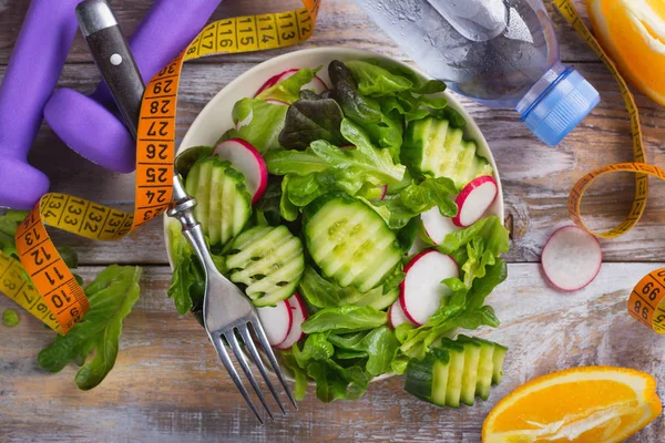 Здоровый вегетарианский салат, гантели и бутылка воды на деревянном столе — стоковое фото