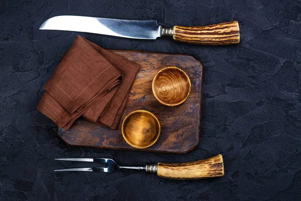 Tabla de cortar de madera, tazón para especias y tenedor vintage y cuchillo sobre fondo negro piedra —  Fotos de Stock