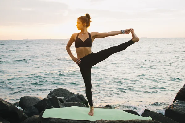 Mujer joven practicando yoga en la costa rocosa al amanecer o al atardecer — Foto de Stock