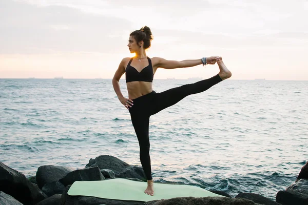 Mujer joven practicando yoga en la costa rocosa al amanecer o al atardecer — Foto de Stock