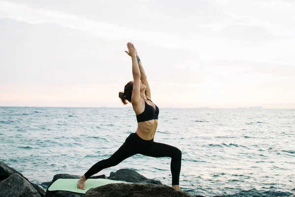 Mujer joven practicando yoga en la costa rocosa al amanecer o al atardecer — Foto de Stock