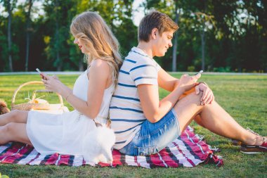 Genç güzel çift parkta oturan ve smarphones kullanarak