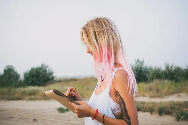 Young hipster woman with pink hair drawing with pencil at the coast — Stock Photo, Image