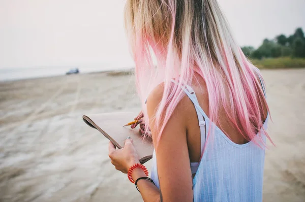 Молода дівчина-хіпстер з рожевим волоссям малює олівцем на узбережжі — стокове фото