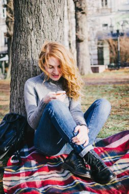 Kızıl saçlı genç kadın kareli ekose parkta oturan ve kullanma telefon bukleler