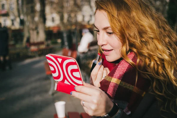 Young beautiful woman sitting  in the park and paints her lips with red lipstick — Stock Photo, Image