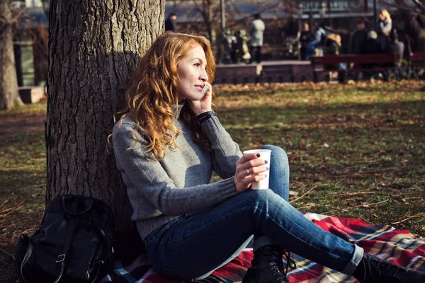 Νεαρή γυναίκα με το ποτήρι του τσαγιού καθισμένος και να μιλήσει στο τηλέφωνο στο πάρκο — Φωτογραφία Αρχείου