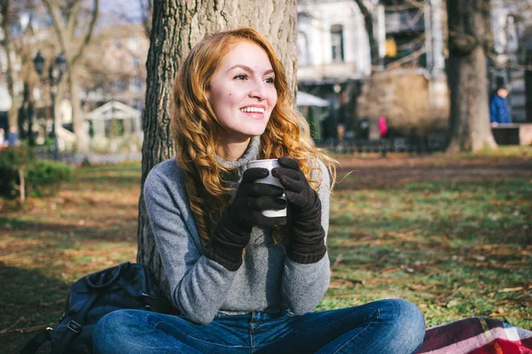 Νεαρή ευτυχισμένη γυναίκα κάθεται με ποτήρι του τσαγιού και χαμογελά στο πάρκο — Φωτογραφία Αρχείου