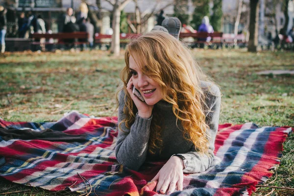 Young woman sitting on plaid and talking on phone in the autumn park — Stock Photo, Image