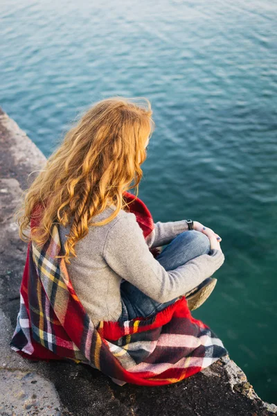 Jeune femme aux cheveux roux boucles assis dans une écharpe au bord de la mer — Photo