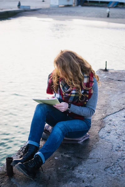 Young woman with red hair curls sitting and drawing with pen at the seacoast — Stock Photo, Image