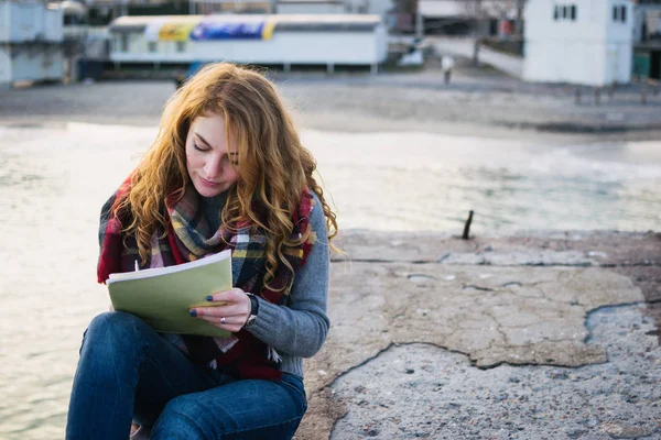 Young woman with red hair curls sitting and drawing with pen at the seacoast — Stock Photo, Image