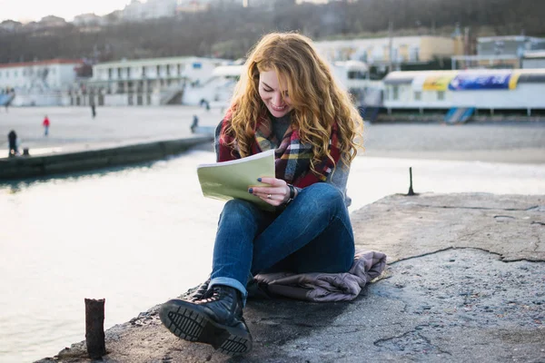 Young woman with red hair curls sitting and drawing with pen at the seacoast — Stock Photo, Image