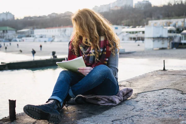 Young woman with red hair curls sitting and drawing with pen at the seacoast — Stock Photo, Image