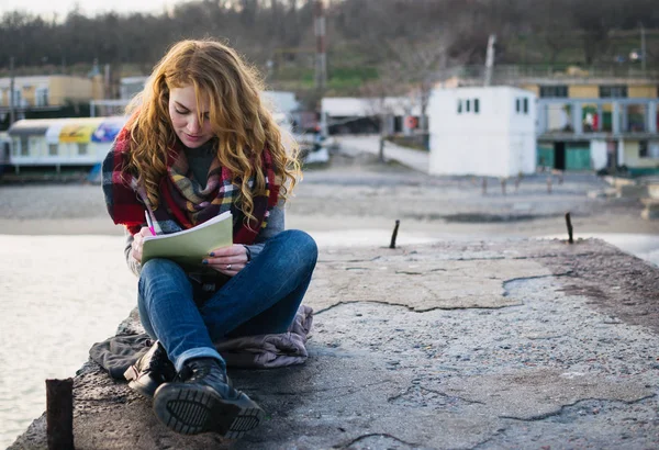 Young woman with red hair curls sitting and drawing with pen at the seacoast — Stock Photo, Image