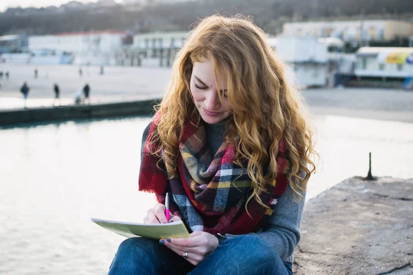 Νεαρή γυναίκα με τα κόκκινα μαλλιά μπούκλες συνεδρίαση και το σχέδιο με πένα σε seacoast — Φωτογραφία Αρχείου