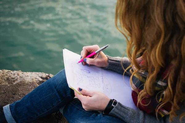 Jovem com cachos de cabelo vermelho sentado e desenho com caneta no litoral, close-up — Fotografia de Stock