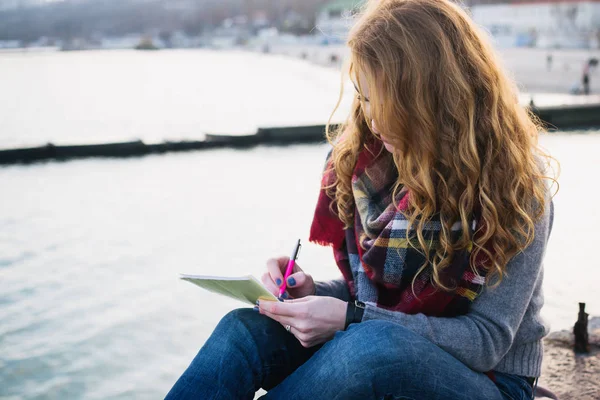 Jovem com cachos de cabelo vermelho sentado e desenho com caneta no litoral — Fotografia de Stock