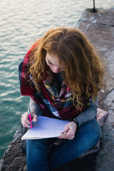 Jovem com cachos de cabelo vermelho sentado e desenho com caneta no litoral — Fotografia de Stock