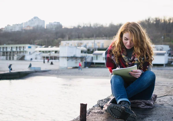 Young woman with red hair curls sitting and drawing with pen at the seacoast — Stock Photo, Image