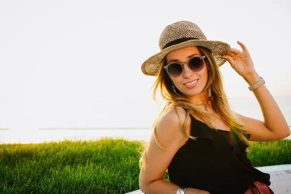 Красива молода жінка в капелюсі і окулярах посміхається і позує на березі моря — стокове фото