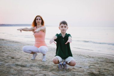 Anne ve oğlu gündoğumu veya günbatımı sırasında deniz sahilinde egzersiz yaparak