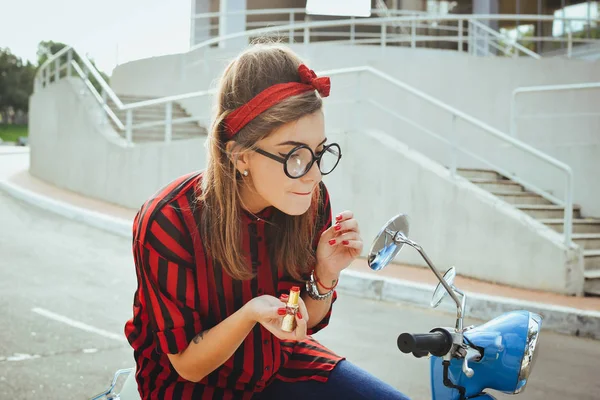 Wanita hipster muda berkacamata duduk di skuter vintage dan mengecat bibirnya dengan lipstik merah — Stok Foto