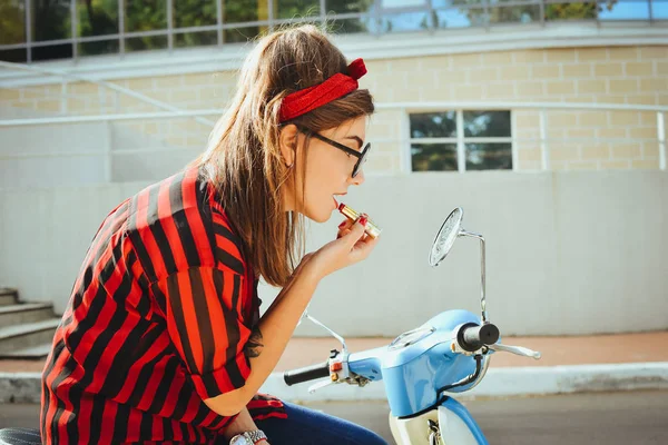 Молода хіпстерка в окулярах сидить на старовинному скутері і малює губи червоною помадою — стокове фото