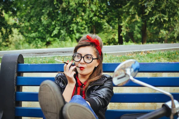 Jonge hipster vrouw in glazen zittend op een bankje en roken sigaret — Stockfoto